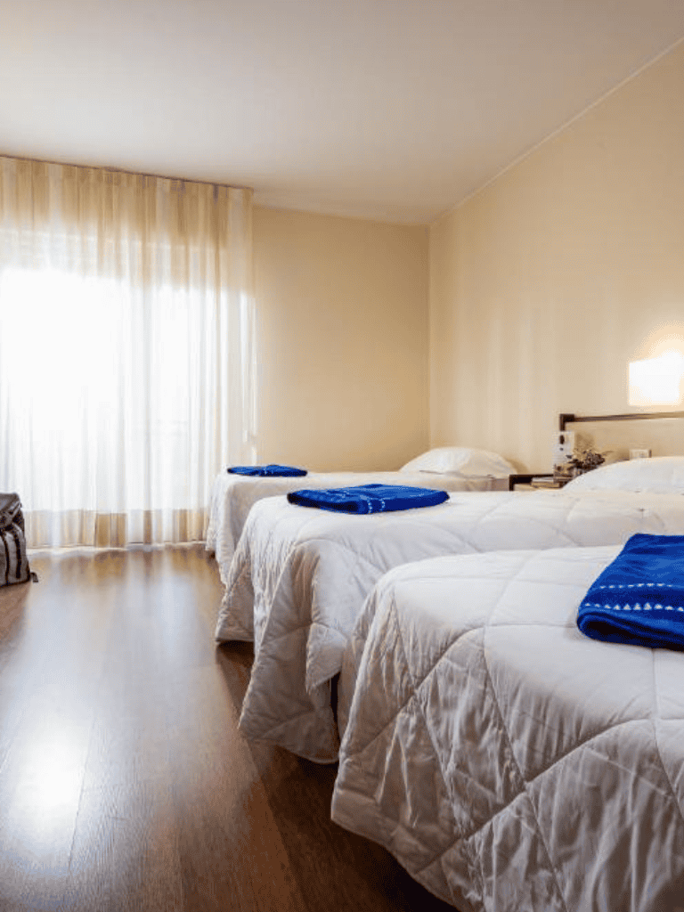 Clean hotel room with three beds and blue towels, bright natural light from window.