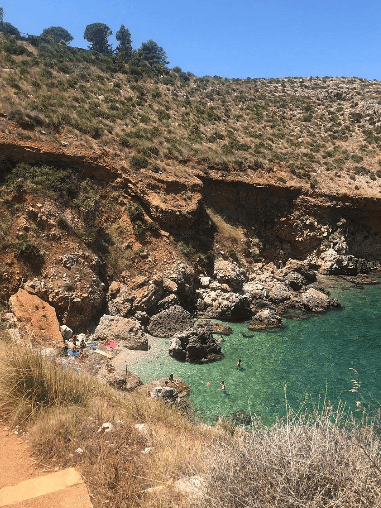 Secluded beach cove with rocky cliffs, clear turquoise water, and people enjoying swimming and relaxing.