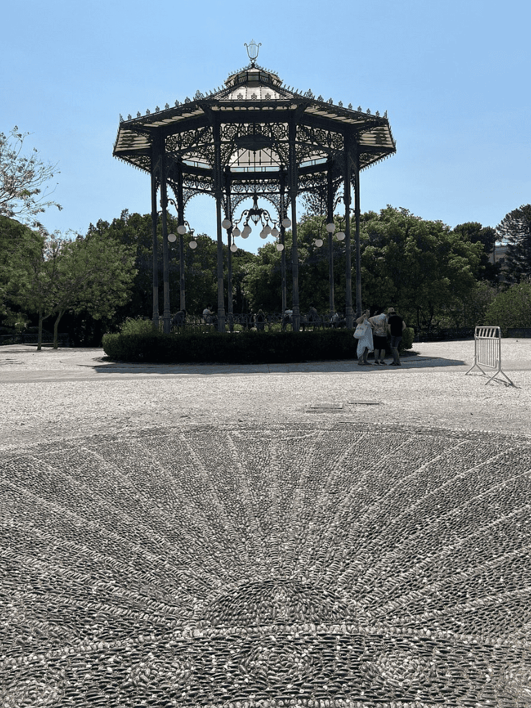 Vintage cast-iron bandstand in a park with a cobblestone sunburst pattern on the ground, surrounded by trees.