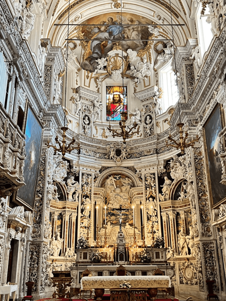 Intricate baroque church altar with ornate white sculptures, gold accents, and religious paintings in a grand historic church interior.