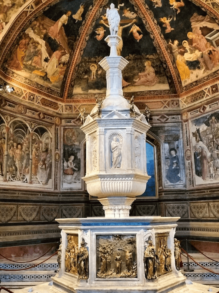 Ornate marble fountain with religious sculptures inside historic church, surrounded by detailed frescoes and artwork.