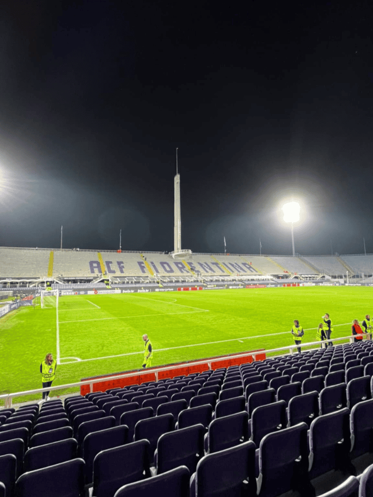 Skyline stadium lit for night football game with security staff on field, empty seats, and "ALAF FIORNIE" on the stands.