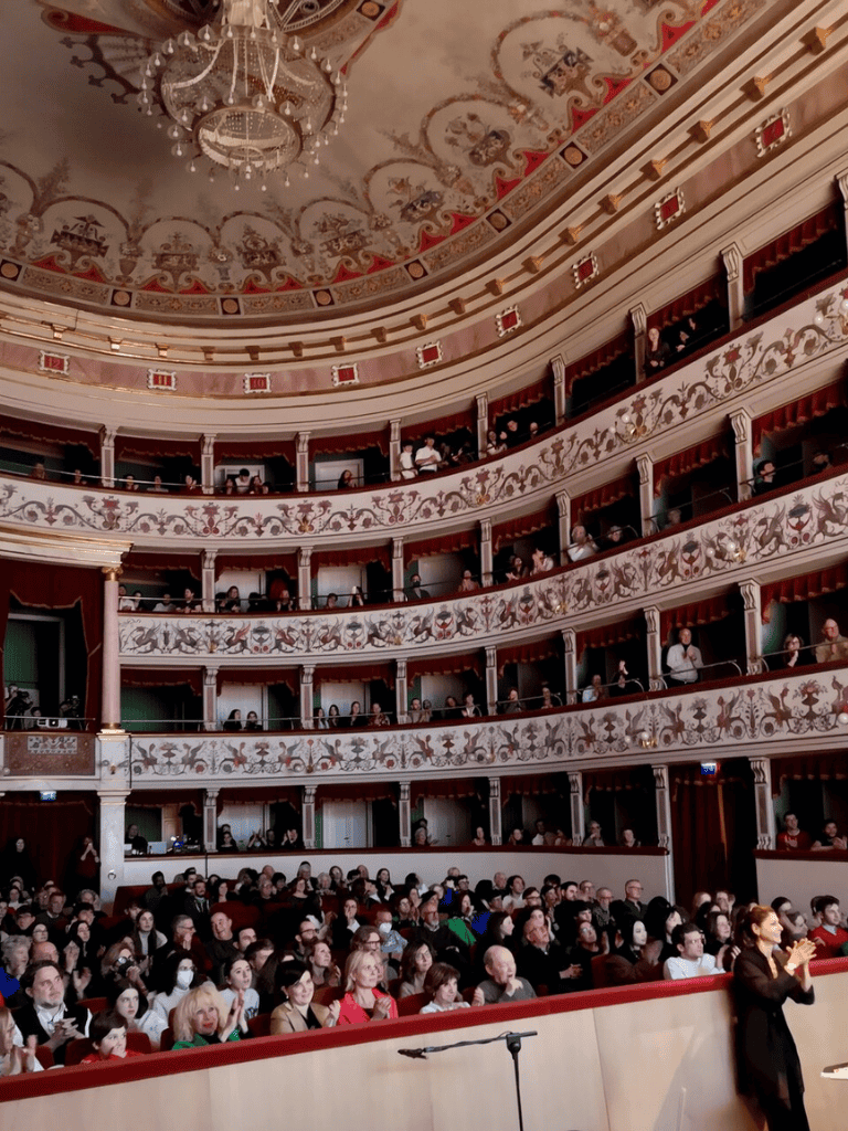 Audience in elegant theater with ornate balcony tiers and chandelier.