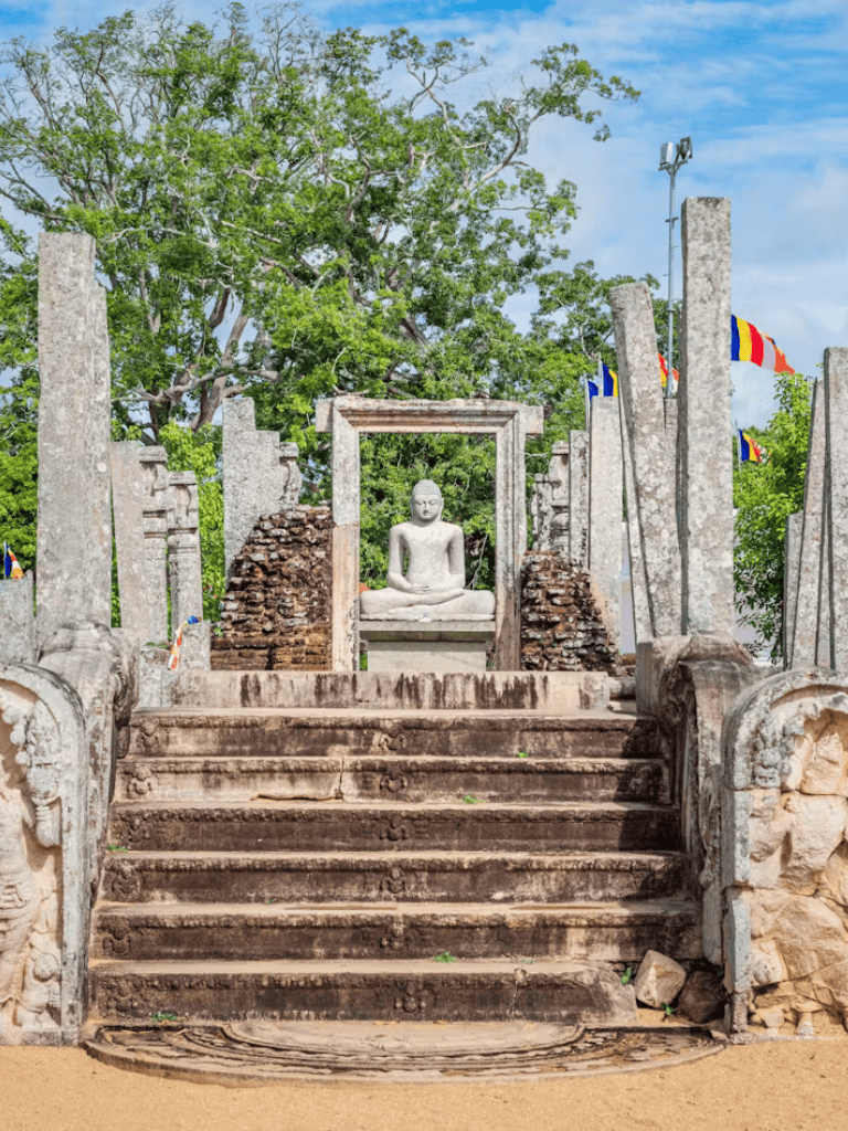 Ancient Buddhist shrine with a meditating Buddha statue and historic stone steps, surrounded by lush greenery and colorful flags.