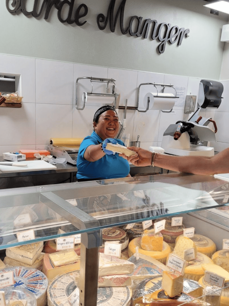 Smiling cheese shop employee offering cheese, variety of gourmet cheeses displayed, friendly customer service experience.