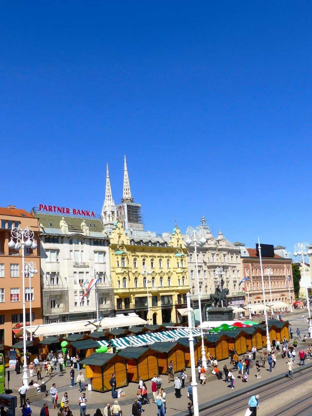 Vibrant city square in Budapest with historic architecture, outdoor markets, and bustling crowds under a clear blue sky.