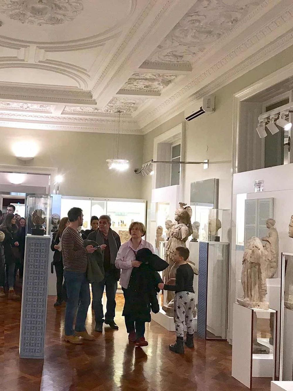 Ancient Greek sculptures displayed in a museum with visitors exploring the collection.