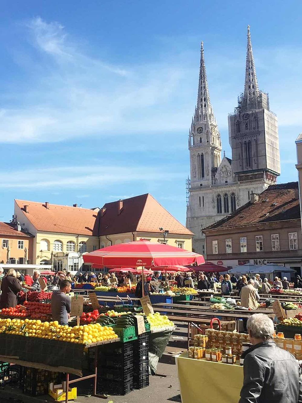 Colorful outdoor market with fresh produce, historic buildings, and a prominent cathedral in the background.