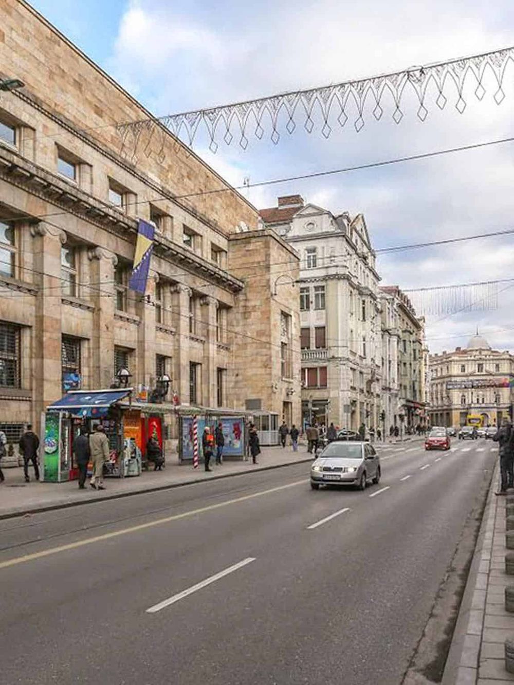 Historic city street with European architecture and traffic, festive string lights overhead.