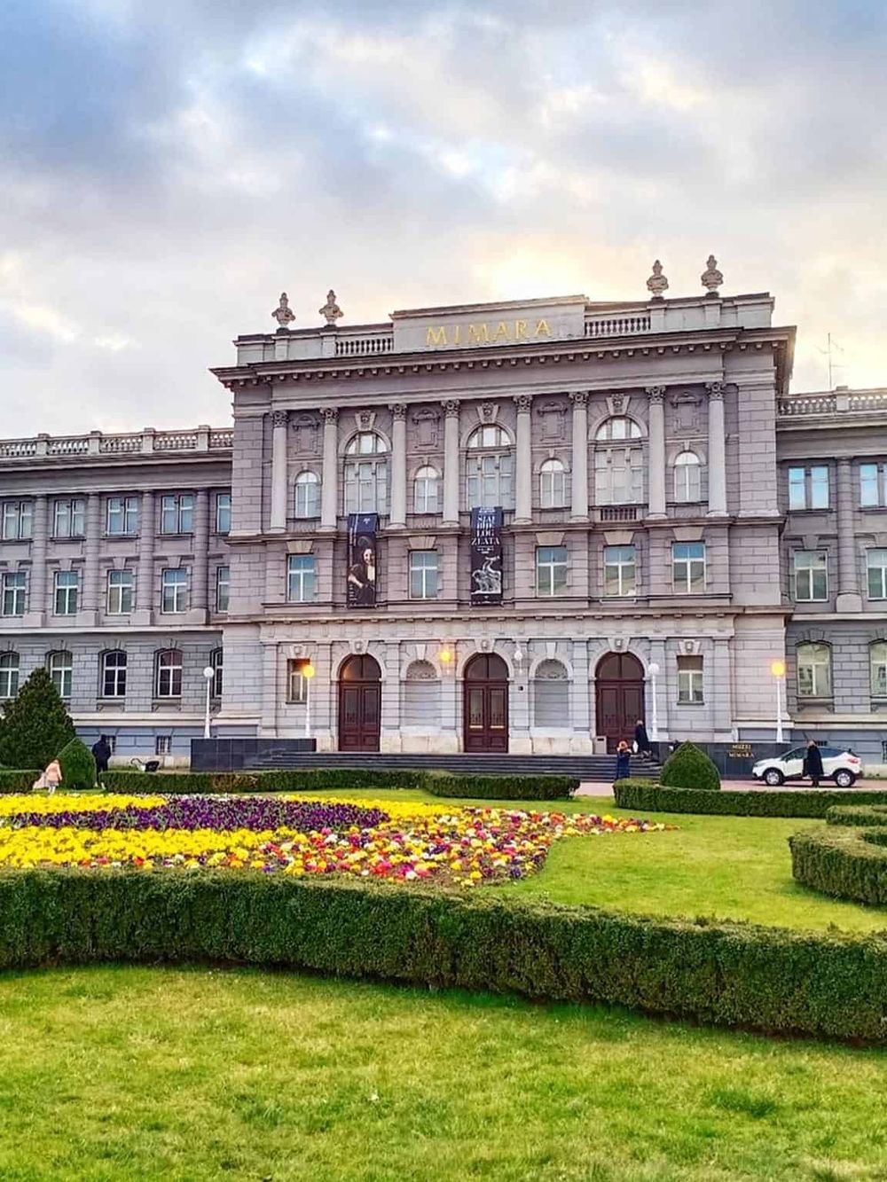 Historic hotel building in a cityscape with colorful flower bed in front and cloudy sky above.