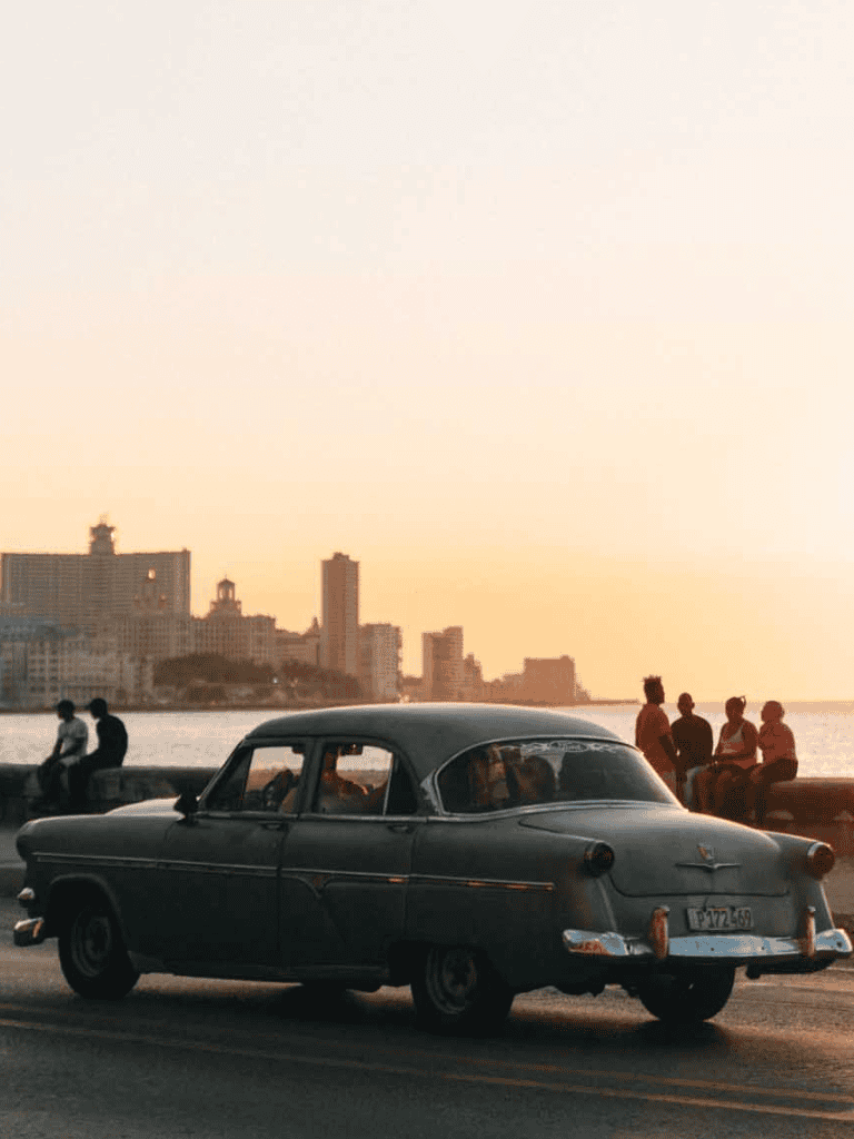 Vintage car cruising along a coastal city at sunset with people enjoying the view.