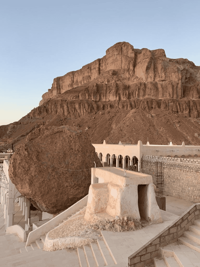 Huge desert rock formation near white building, desert landscape, sunny clear sky.