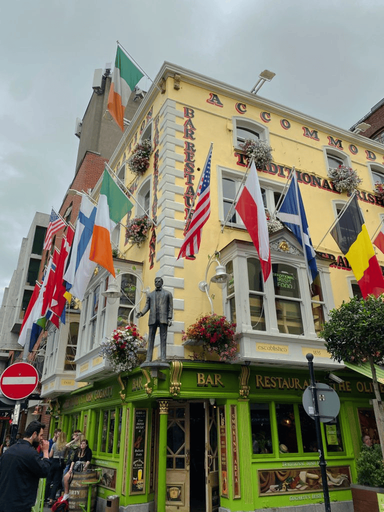Irish pub with colorful flags, flowers, and a statue of a man outside, located on a lively city corner.