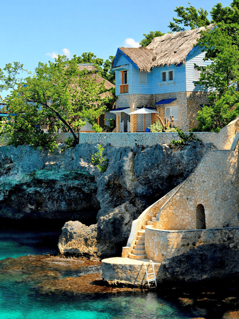 Coastal seaside house with stone steps and tropical greenery on rocky shore.