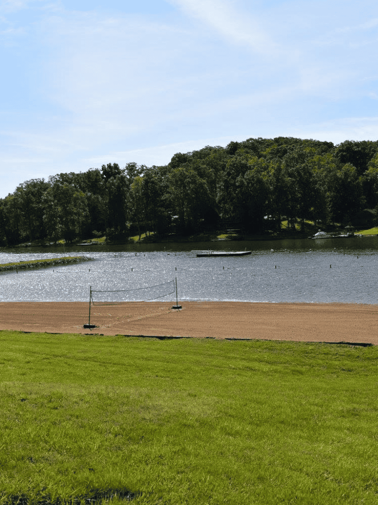 Serene lakeside scene with sandy beach, lush green grass, and trees in the background.