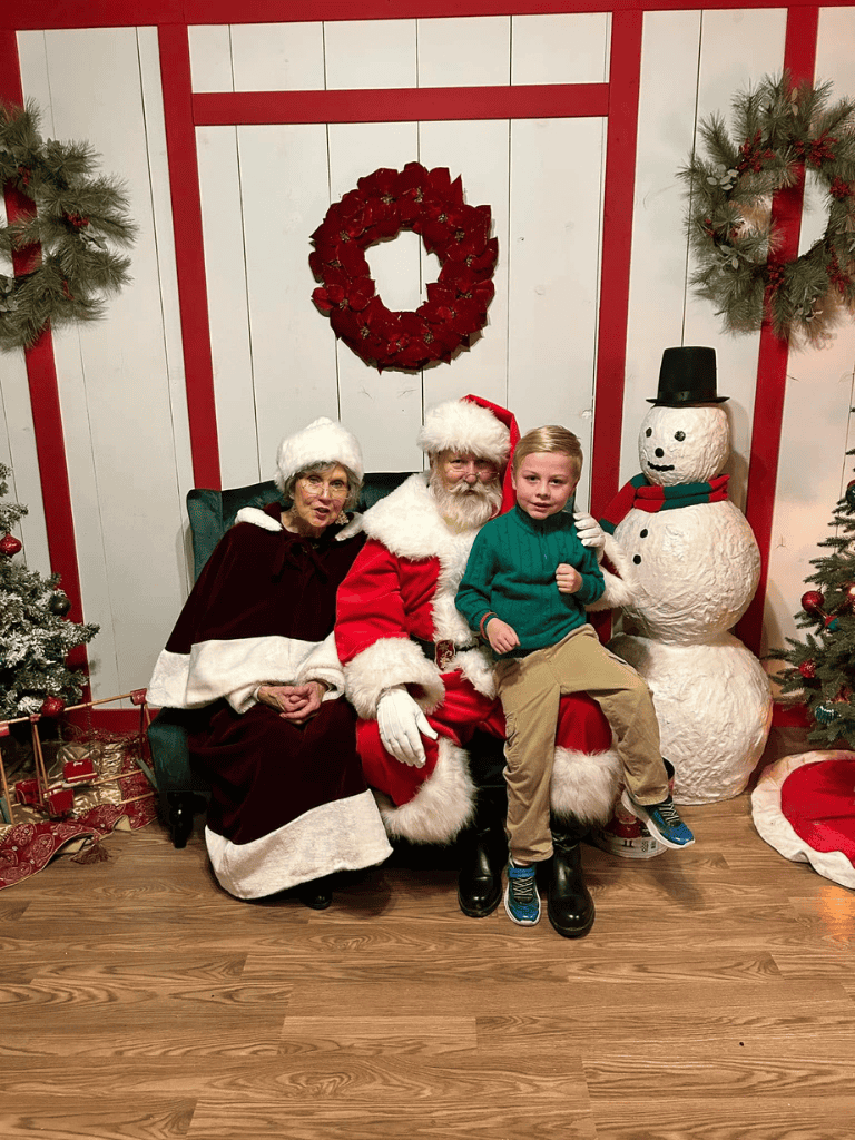 Santa Claus with Mrs. Claus and a young boy at a Christmas themed photo op featuring holiday decor and a snowman.