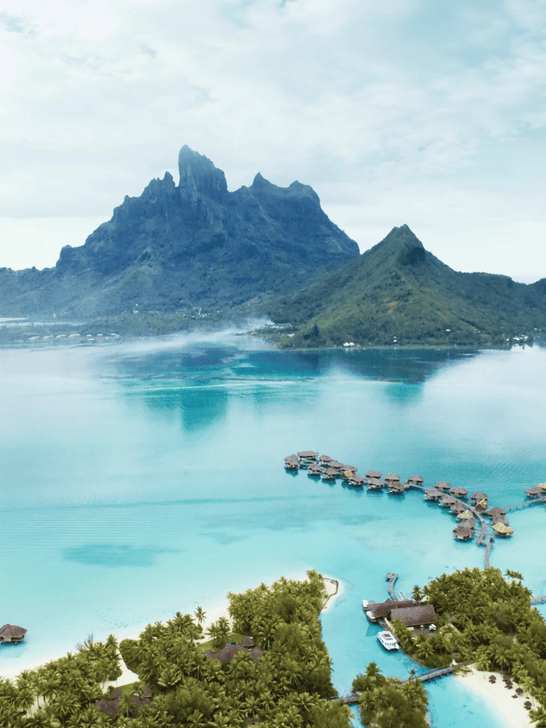 Tropical island resort with overwater bungalows beneath lush mountains and clear blue water.