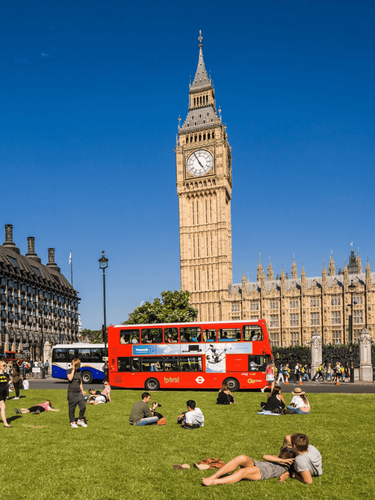 Double-decker red bus in front of Big Ben in London, UK, with tourists and locals enjoying the sunny day.