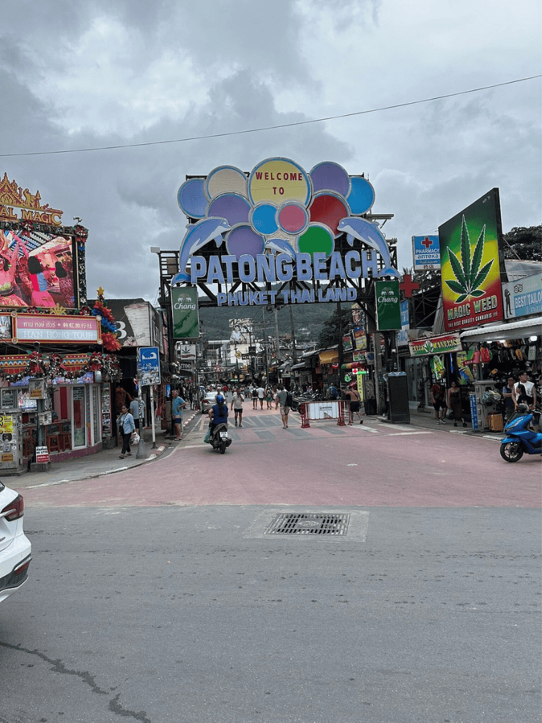 Colorful Patong Beach entrance with crowd in Phuket, Thailand, featuring tourist shops and neon signs.