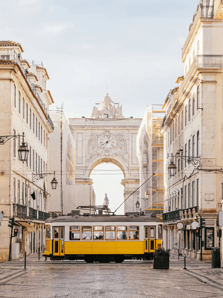 Yellow Lisbon tram on historic city street with arch and clock tower in background.