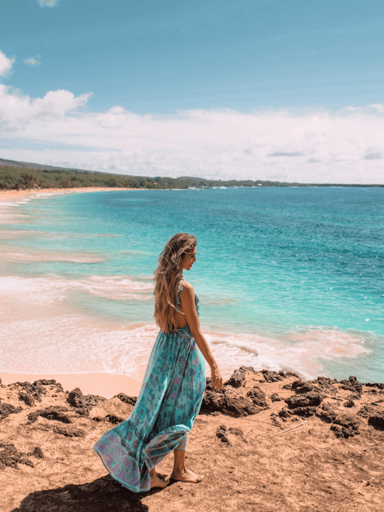 Serene woman walking along tropical beach with turquoise waters and rocky shoreline, scenic seaside vacation scene.