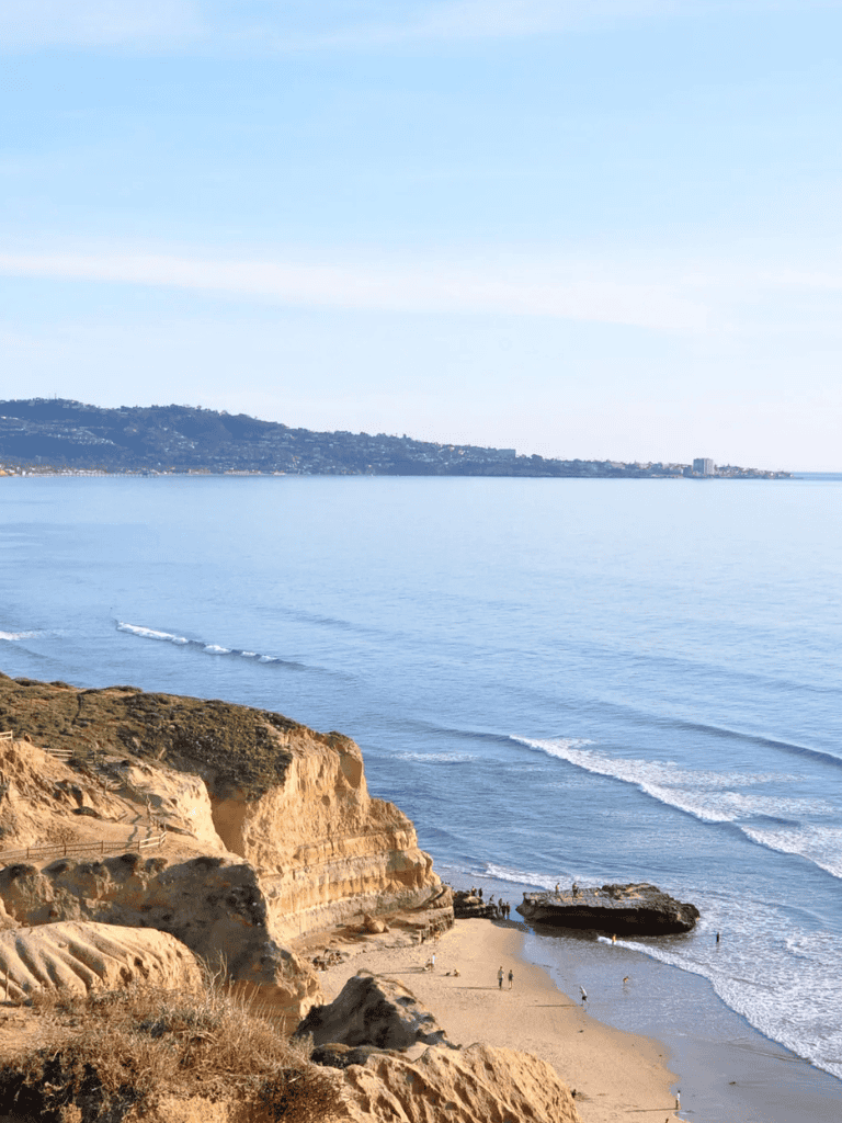 Seaside cliffs overlooking a calm ocean with people on the beach and distant shoreline view.