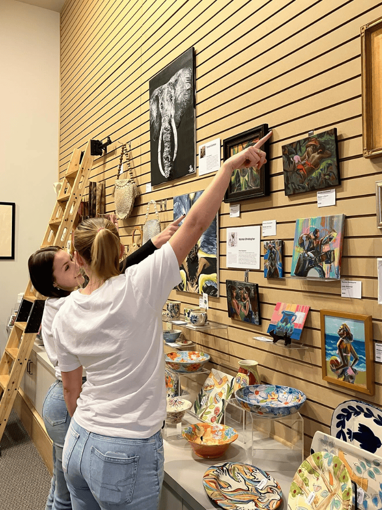 Colorful art gallery with pottery displays and visitors examining paintings on a wooden wall.