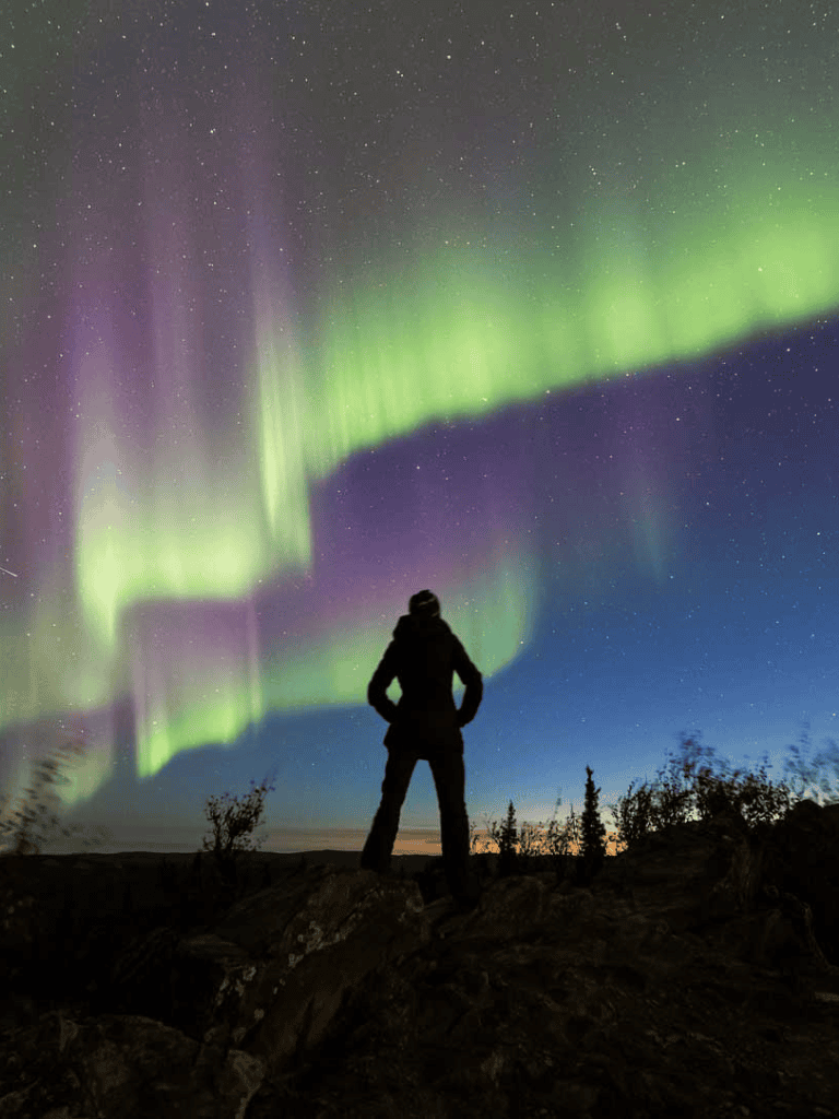 Aurora Borealis over person standing on rocky terrain, Northern Lights display in the night sky.