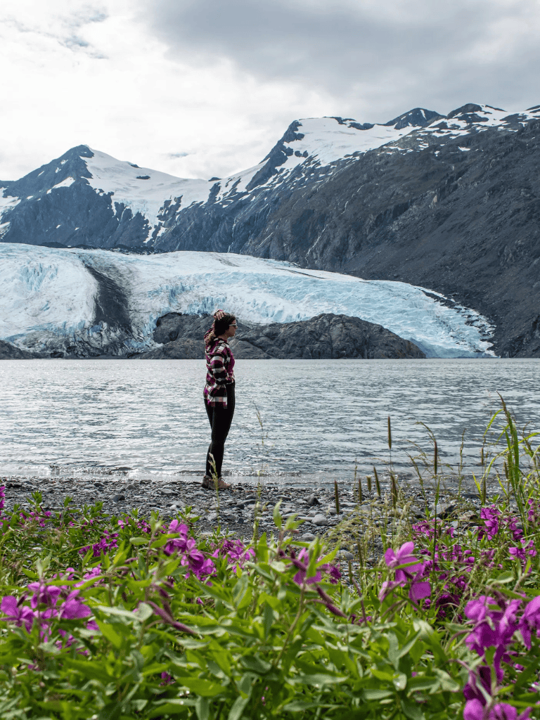 Snow-capped mountains and glacier in Alaska with a woman standing by a lake, exploring adventure travel destinations.