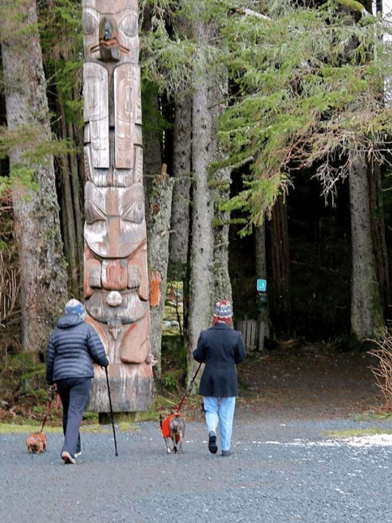 Colorful totem pole in lush forest with people walking dogs, showcasing outdoor adventure and cultural landmark in nature.