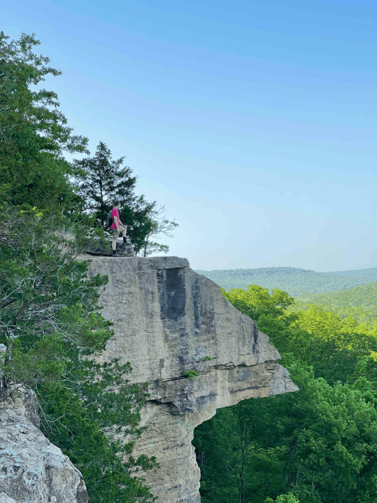 Climber on a rock ledge overlooking lush green forest with mountains in distance.