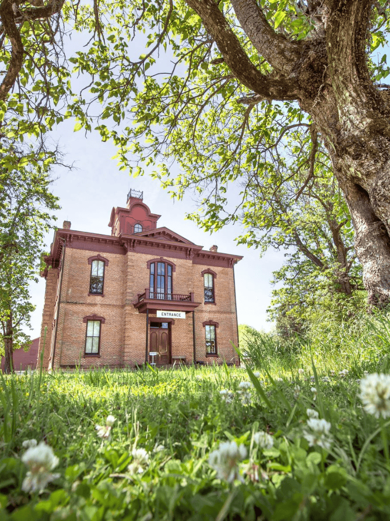 Old historic brick building with entrance sign, lush green trees and grass, bright sunny day.