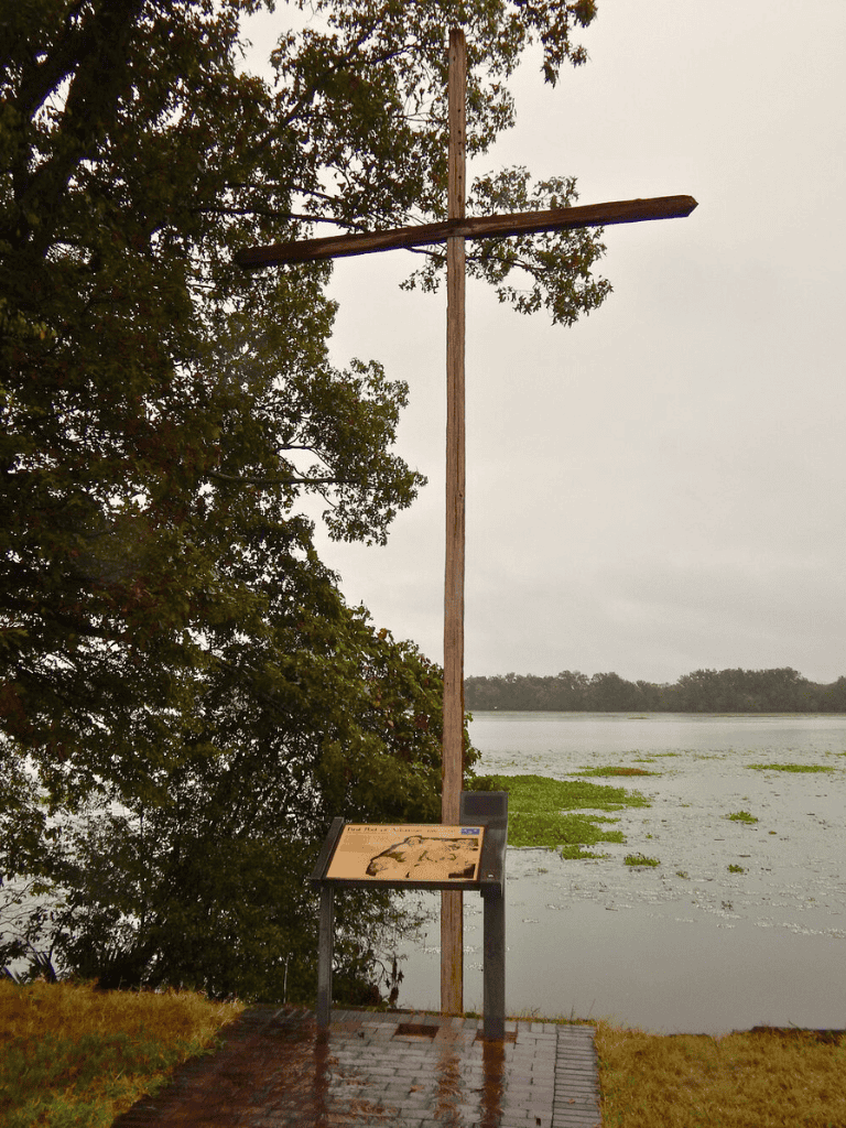 Old wooden directional cross by lake, surrounded by trees, with informational sign in front, during overcast day.