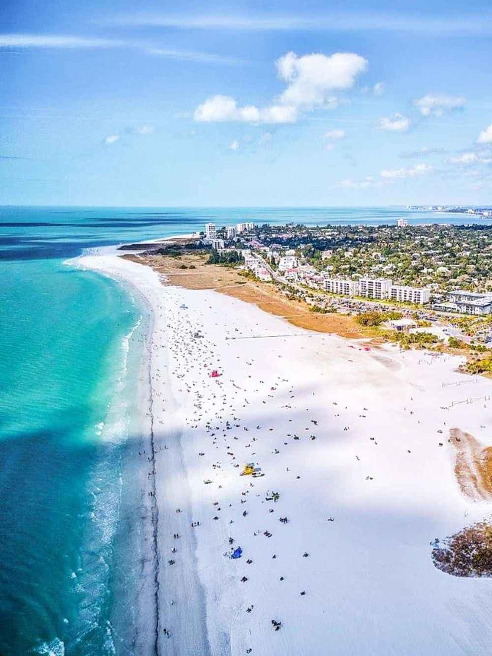 Breathtaking aerial view of a sunny beach with turquoise waters, white sands, and a lively coastal cityscape.