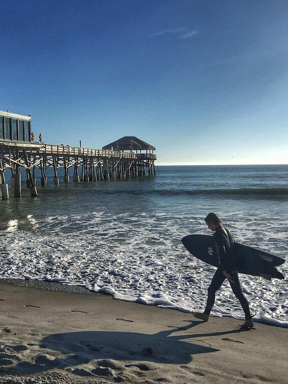 Surfer walking on sandy beach with a surfboard under a sunny sky at a pier.