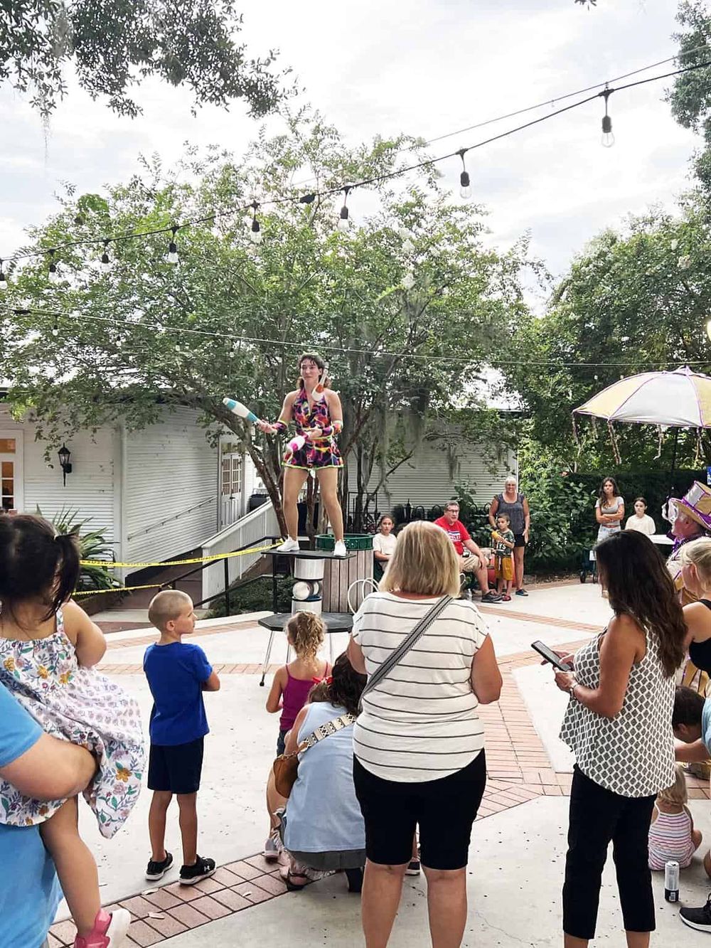 Acrobat performing a balancing act at an outdoor event with children and adults watching under string lights.