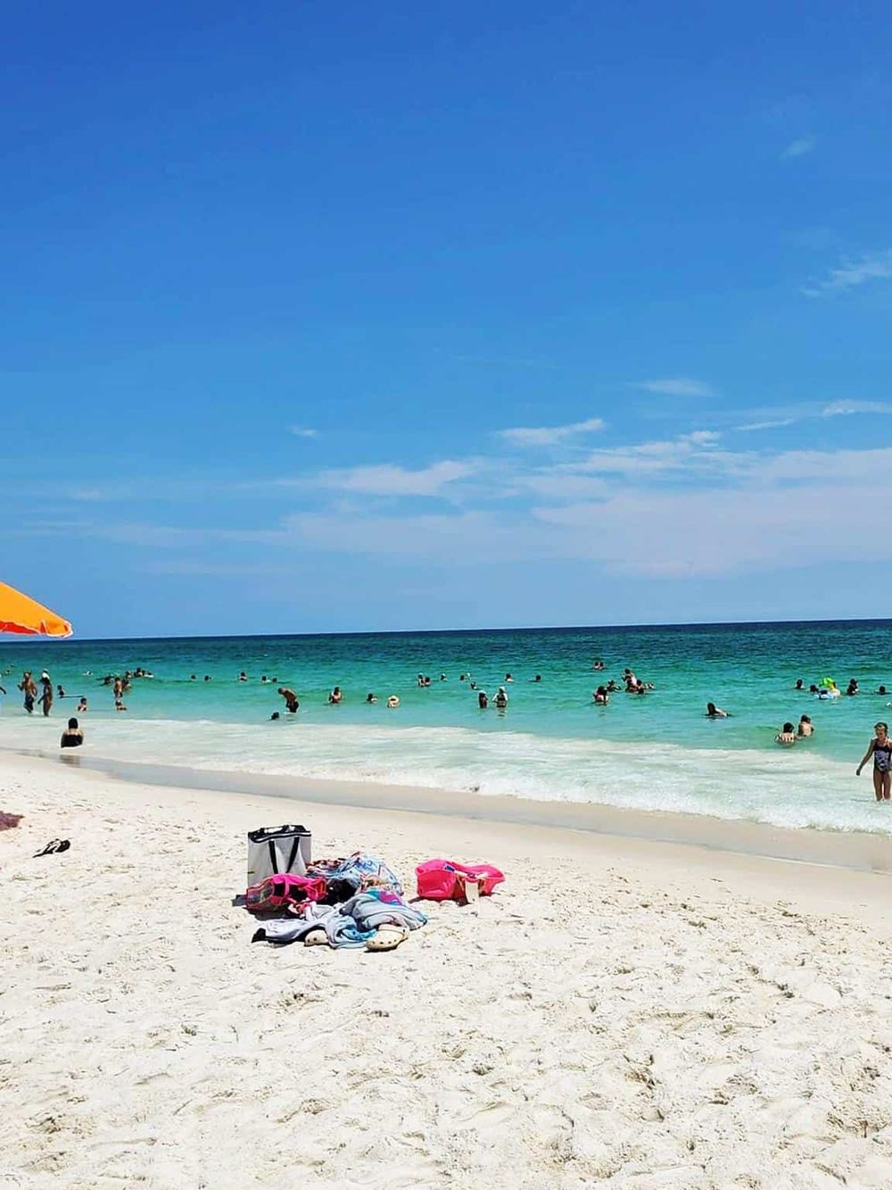 Bright beach scene with sunbathers and swimmers, clear blue sky, and turquoise ocean waves.