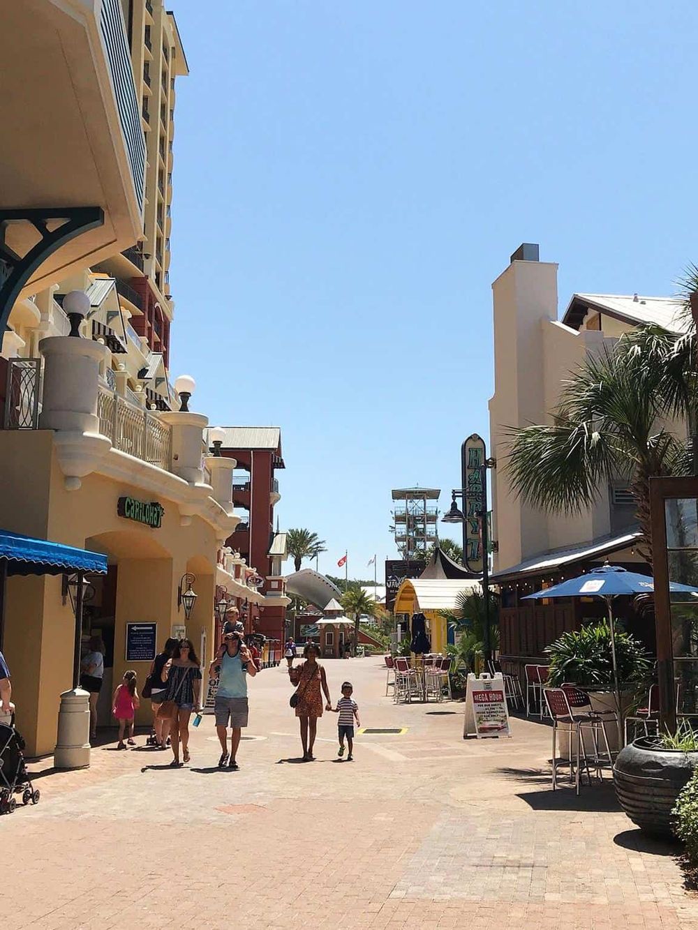 Bright outdoor shopping district with shops, palm trees, and families enjoying sunny weather.