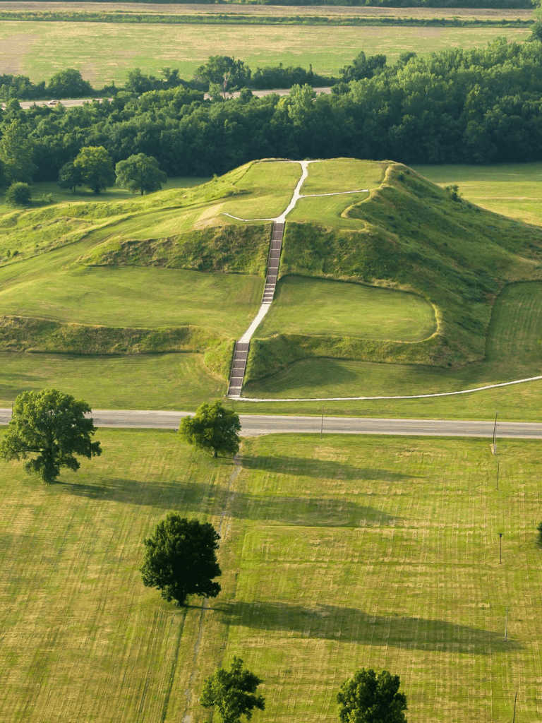 Ancient mound surrounded by lush green landscape and trees, historical earthwork site, scenic rural area.