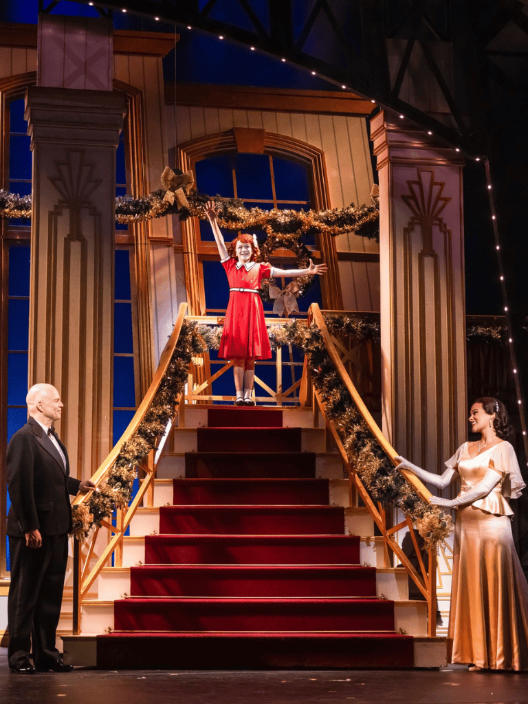 Cheerful holiday musical theater scene with performers on festive staircase backdrop, Christmas decorations, and joyful atmosphere.