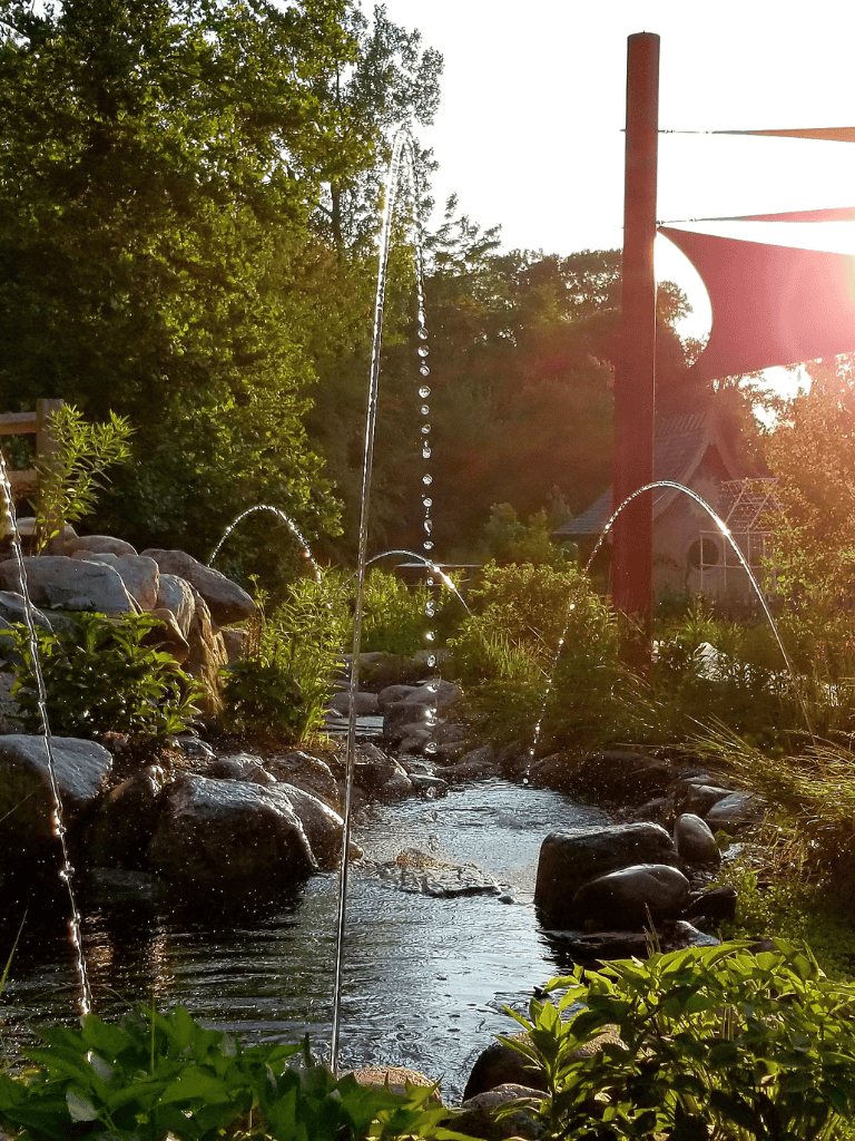 Tranquil garden stream with fountain, rocks, and lush greenery at sunset, perfect for outdoor relaxation.