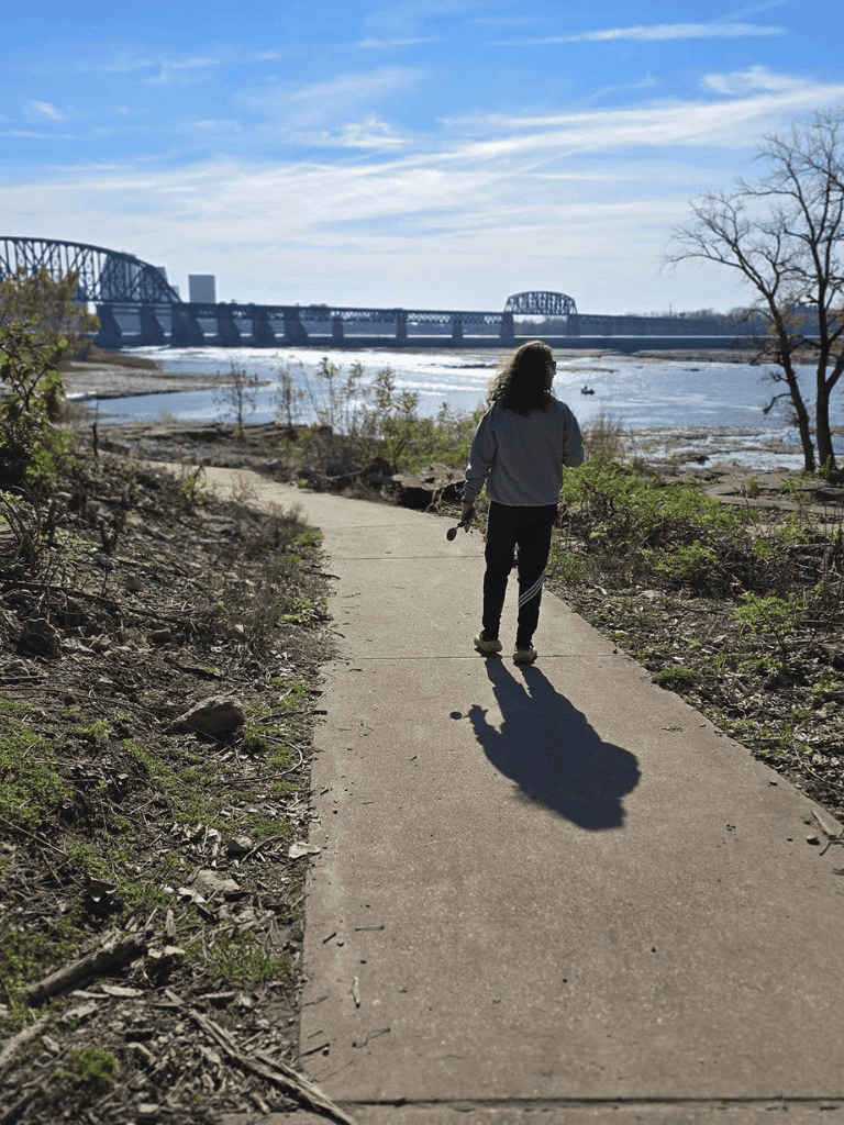 Peaceful riverwalk scene with a woman walking along a narrow path with a bridge in the background, inspiring outdoor exploration.