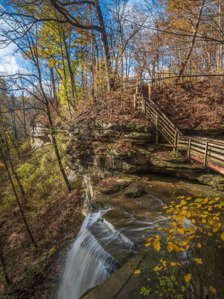 Waterfall and hiking trail on a forested hillside with stairs and railings in autumn, scenic outdoor adventure.
