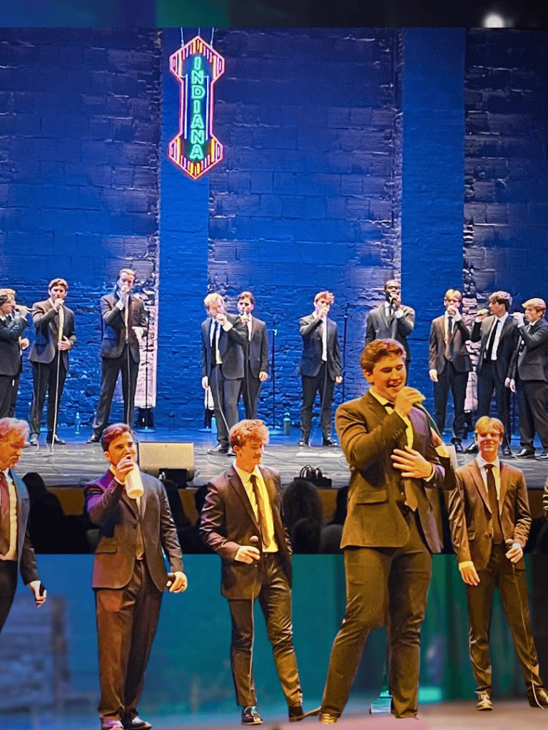 Young male singers performing on stage at a musical concert, in formal suits, with vibrant stage lighting and a neon church sign backdrop.