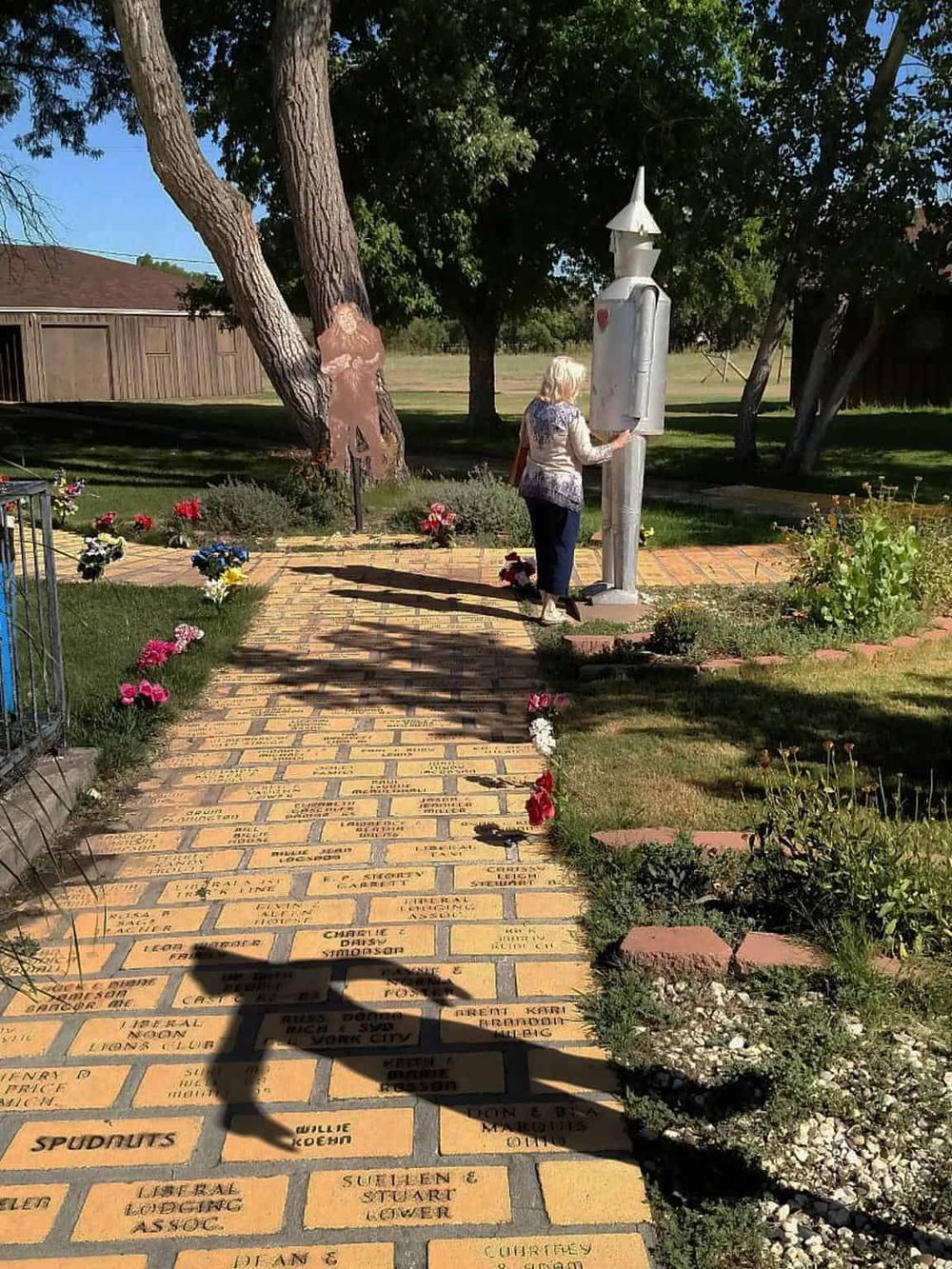 A woman reading a memorial plaque at the QuestForDirections site with a paved walkway, flowers, and trees.