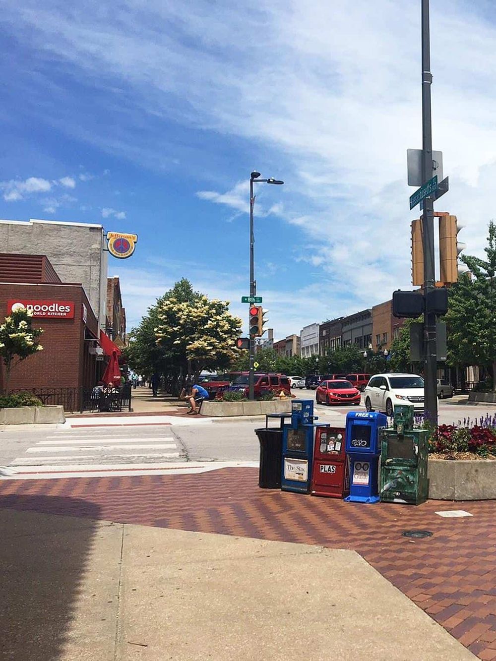 Colorful city street with newspaper stands, trees, pedestrians, and traffic lights for urban navigation.