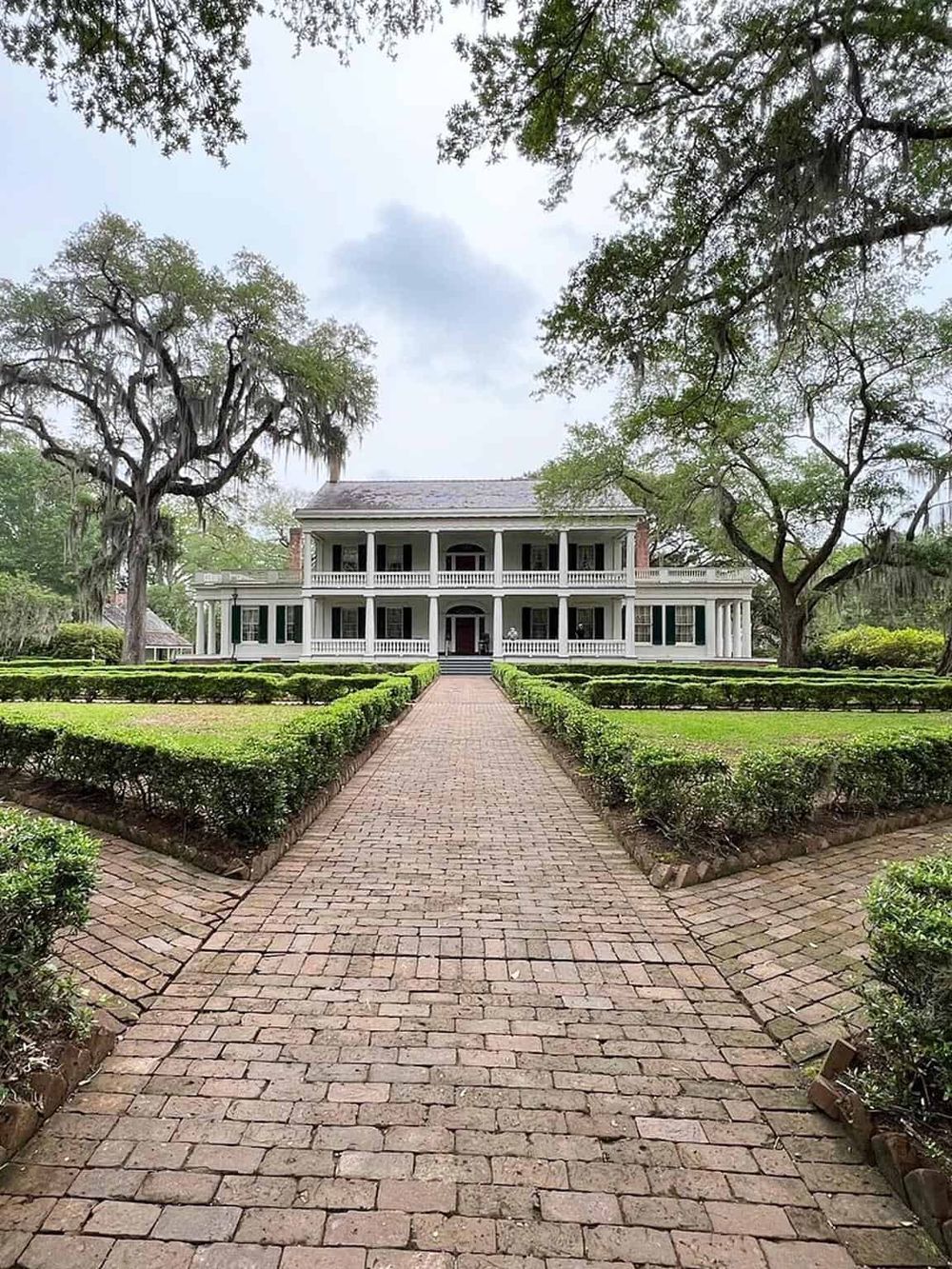 Elegant historic mansion with manicured gardens and brick pathway in a lush, tree-filled setting.