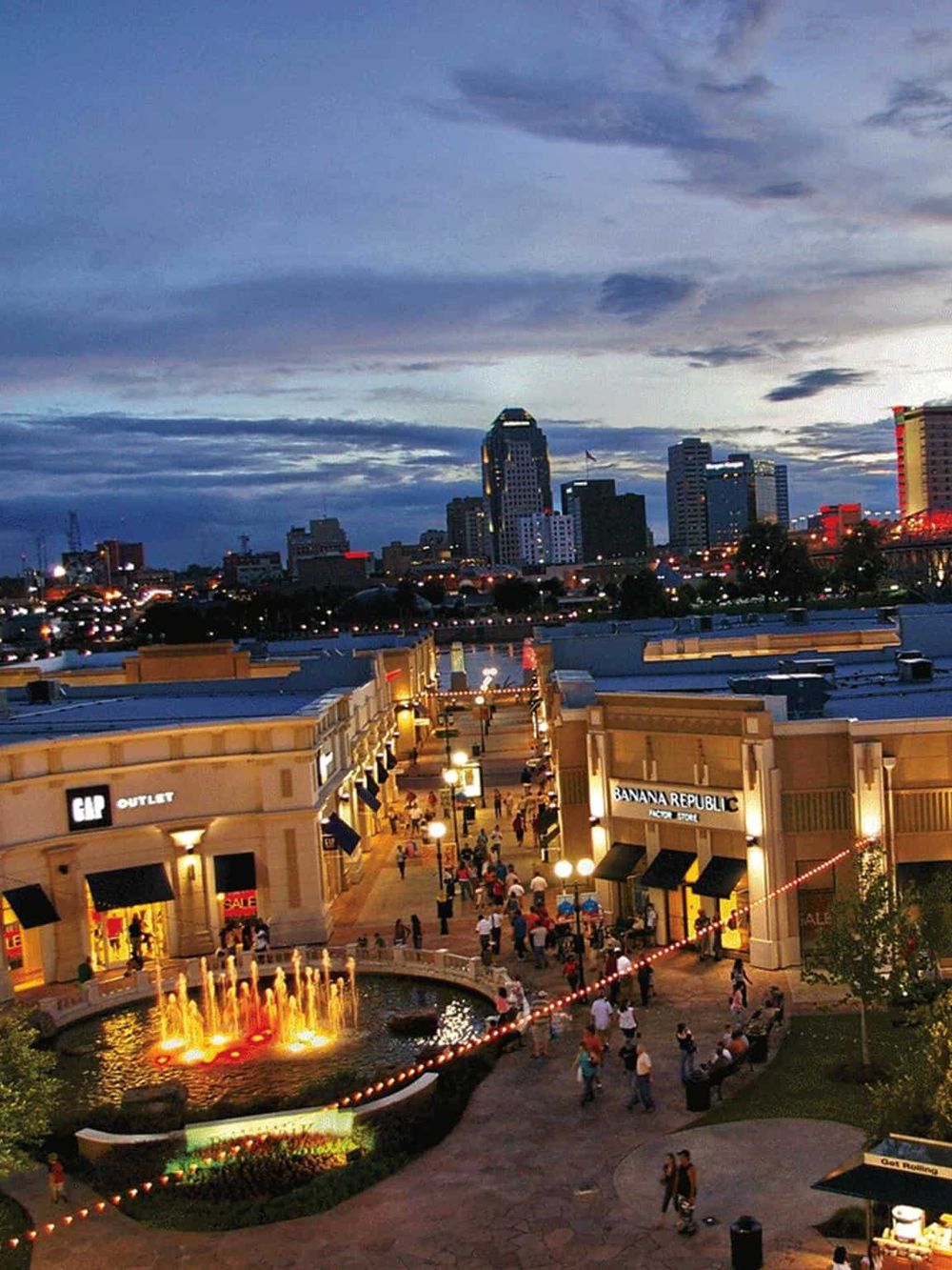 Vibrant outdoor shopping mall with city skyline at dusk, illuminated fountains, and busy shoppers for urban lifestyle enjoyments.