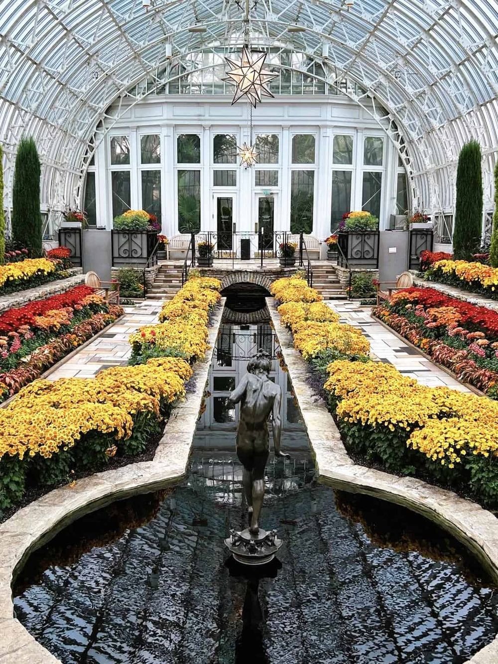 Colorful floral conservatory with glass ceiling, water fountain, and sculpture for garden inspiration.