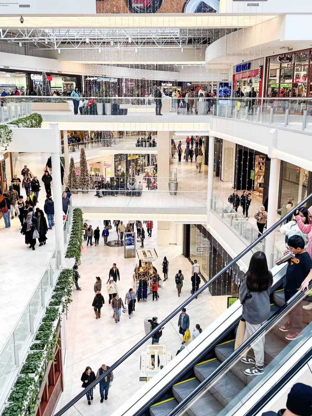 Bright indoor shopping mall with multiple floors, escalators, and shoppers exploring retail stores and festive holiday decorations.